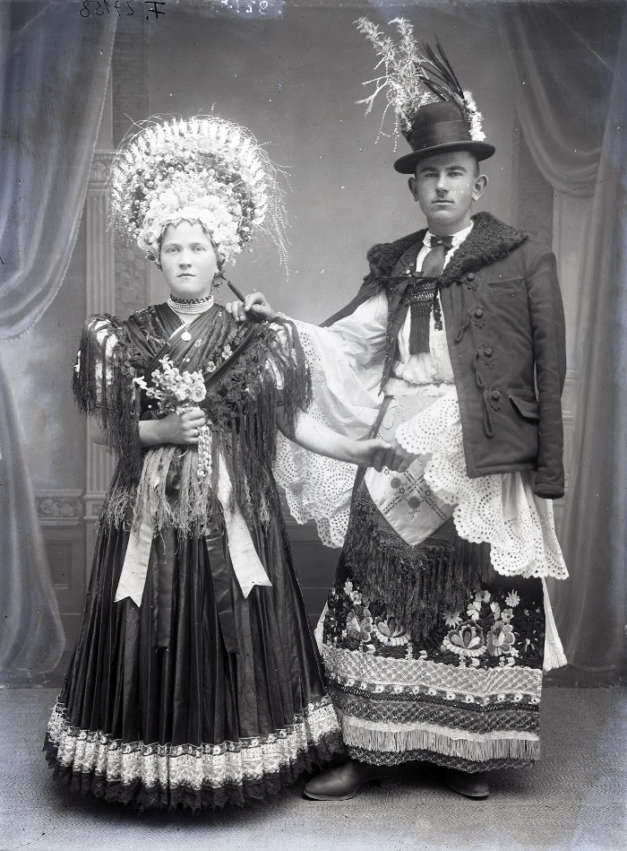 Gáspár József és menyasszonya. A menyasszony fekete esküvői ruhában, színes koszorú a fején, fekete selyem vállkendőben. A vőlegény körtés mintáj fehér lyukhímzéses lobogó ujjú ingben és surcban, barcikalapban, rokkal, Mezőkövesd, üvegnegatív, 18x24 cm, Weissbach Rilly és testvére, 1910-es évek második fele, Néprajzi Múzeum, F 27158