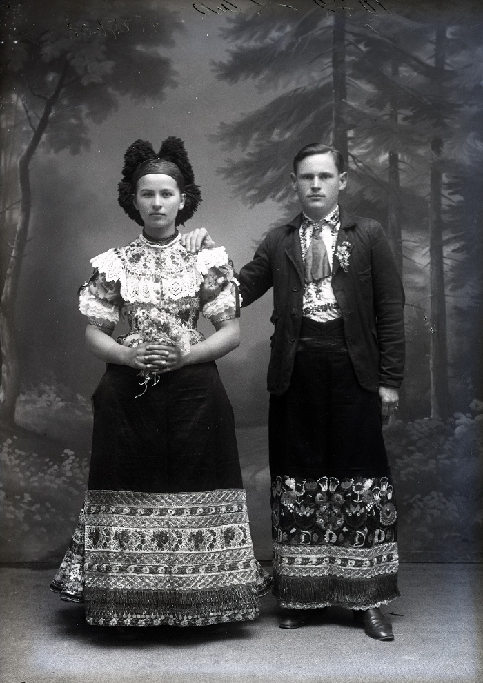 Fiatal házaspár, sajátos változatot mutató öltözetben: az asszony csavarítós kendőben, ráncikában, ragyogóval díszített pántlikás kötőben, a férfi felöltőben, szabott ingben, díszes ingmellben és ragyogóval díszített srucban, Mezőkövesd, üvegnegatív, 12x16,5 cm, Weissbach Rilly és testvére, 1910-es évek második fele, Néprajzi Múzeum, F 27608